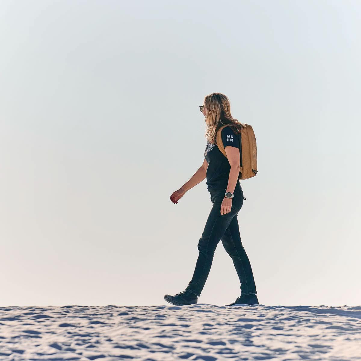 Woman rucking with tan GORUCK backpack on sandy terrain under clear sky