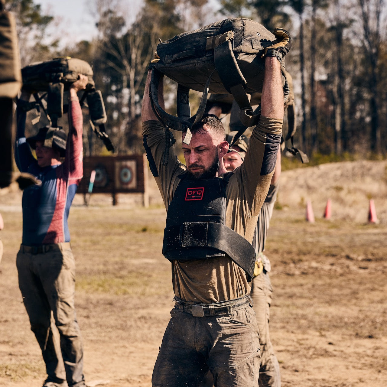Three people lift heavy sandbags overhead while using the Rucker 5.0 during an outdoor rucking fitness challenge on a dirt field.