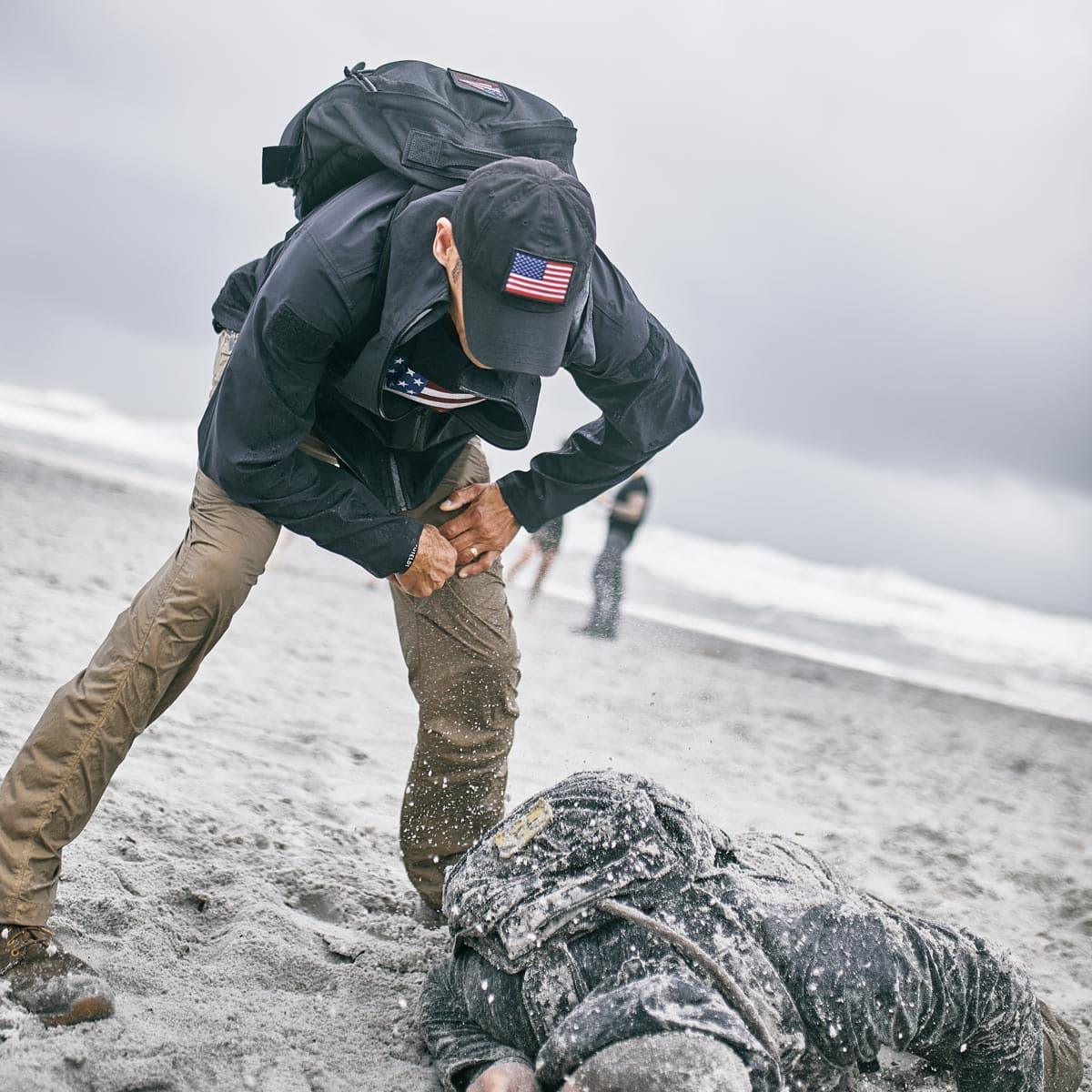 Man in GORUCK gear rucking on a sandy beach, helping teammate during outdoor training event