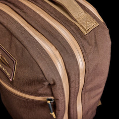 Close-up of the Double Bullet Ruck w/ Bombproof Laptop Compartment - 18L: a brown travel backpack with tan zippers, bombproof laptop storage, and a small patch on the front pocket.