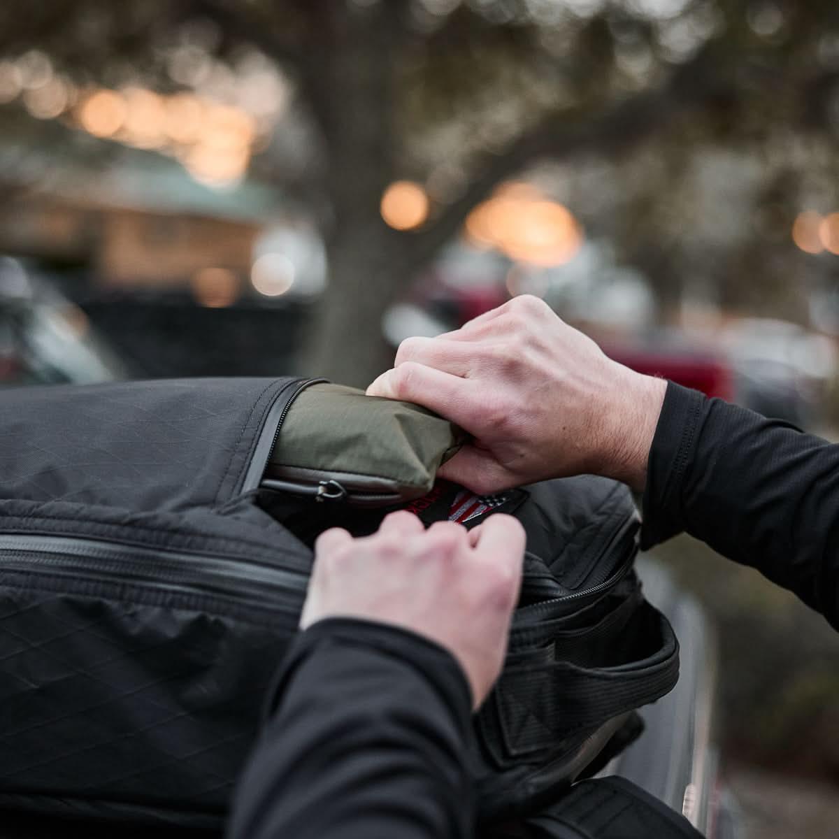 Someone packs the Packable Bullet Ruck - Ripstop ROBIC® into a lightweight backpack while outdoors at sunset.