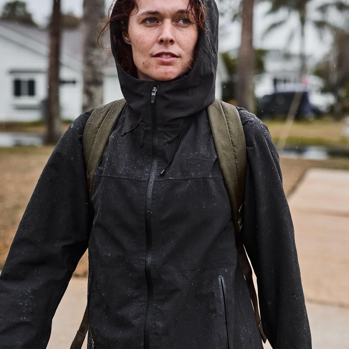 A person carries the Packable Bullet Ruck - Ripstop ROBIC® backpack while walking outside in the rain, wearing a black rain jacket.