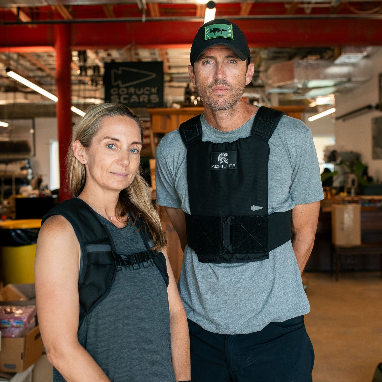 Joe and another person wear Achilles Vest weighted vests while standing together indoors in a gym, surrounded by various fitness equipment.
