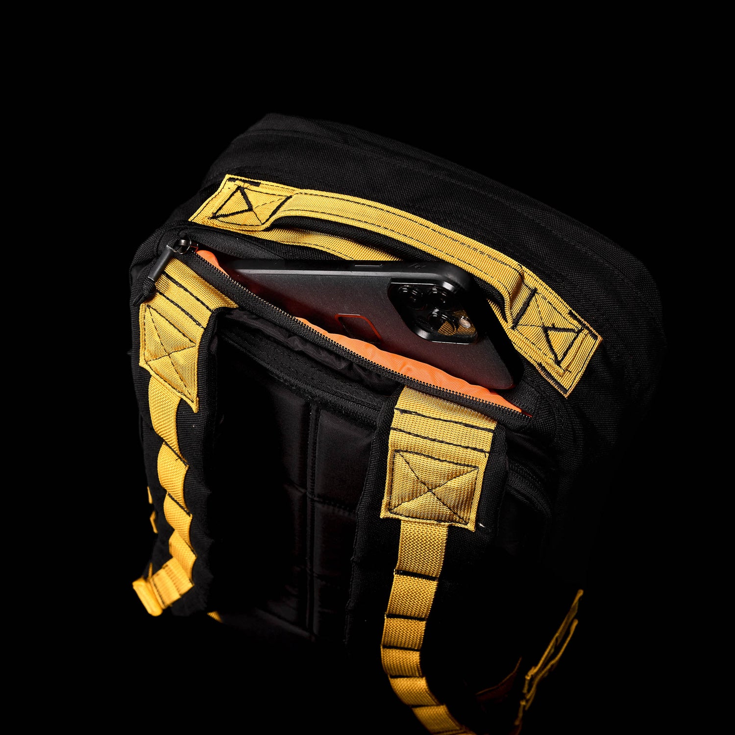A smartphone is partially visible in the zippered pocket of a black and yellow GR1 backpack, crafted from Special Forces grade materials, against a black background.