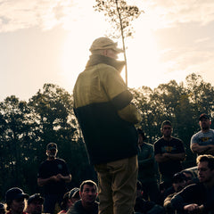 A man wearing the Men’s Task Force Dagger Jacket - Polartec Fleece speaks to a group outdoors at sunset, framed by trees and fading sunlight.