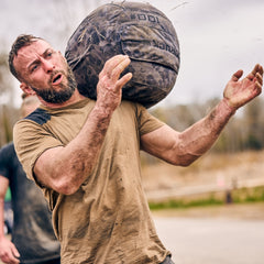 During an intense outdoor fitness challenge, a man lifts the Sand Medicine Ball, showcasing its durability and grit in tough conditions.