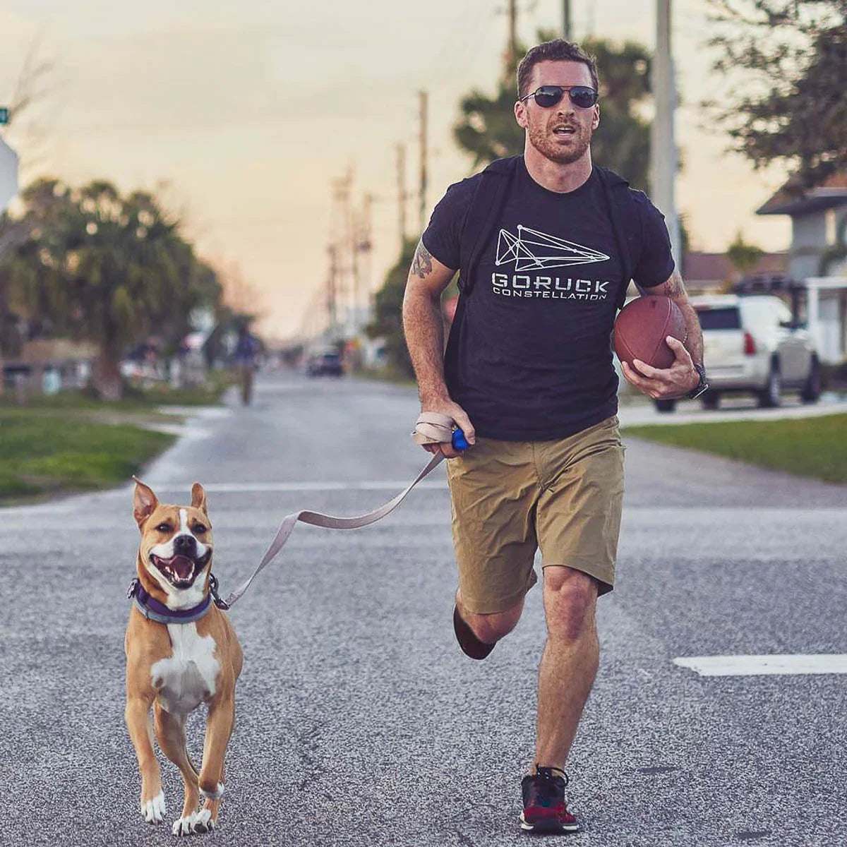 Man in GORUCK shirt and khaki shorts jogging with dog and football on suburban street