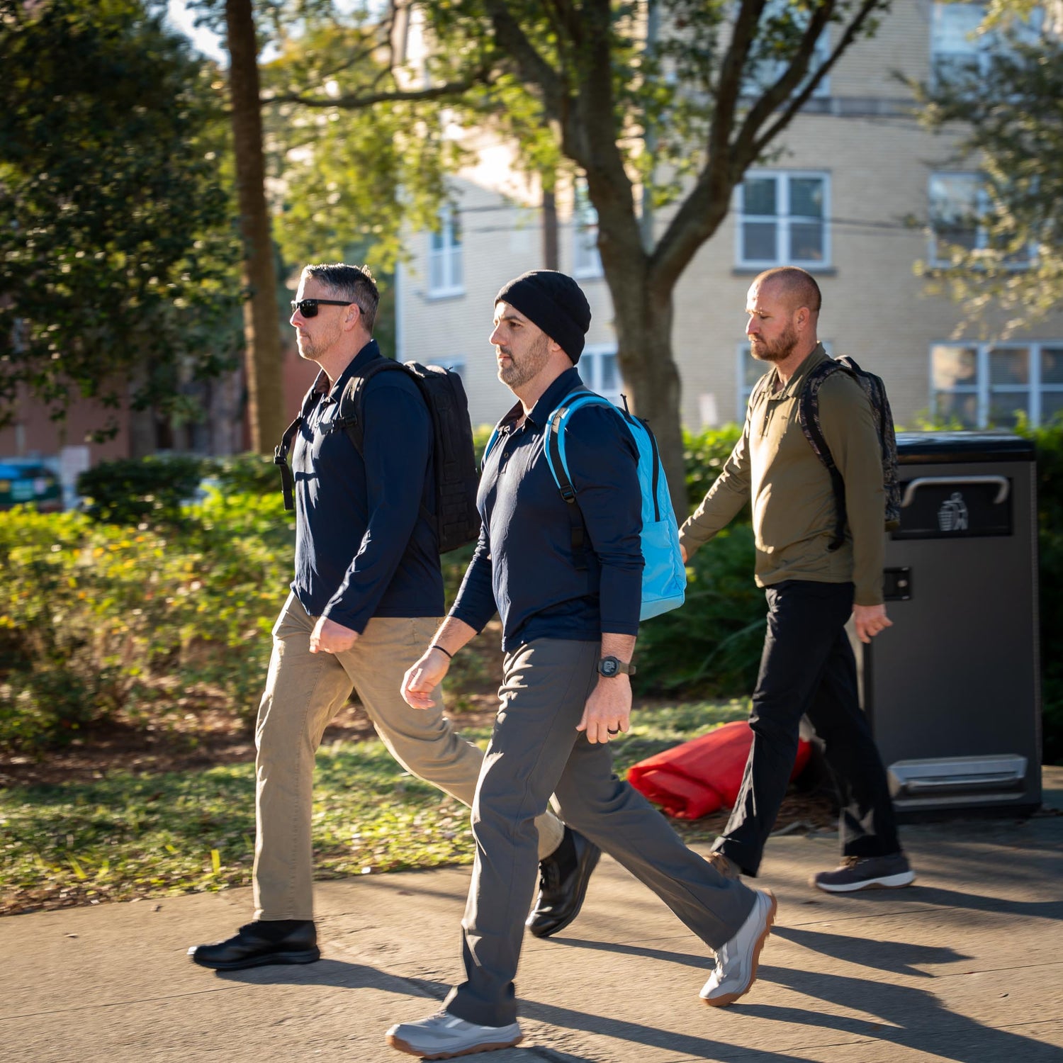 Three men with backpacks walk outside on a sunny day, passing trees and a trash bin—each carrying the Basic Rucker®, ideal for their rucking adventure.
