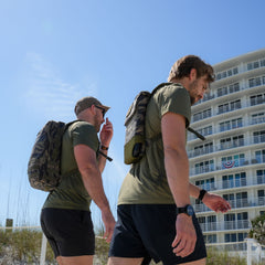 Two men in green shirts and backpacks, including a Ruck Plate Carrier 3.0 with ergonomic lumbar support, walk outdoors near a modern, curved building on a sunny day.