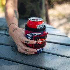 A tattooed hand holds a can of Budweiser in a vibrant Ballistic Beer Jacket - GORUCK koozie made from tough 1000D CORDURA, resting on a wet outdoor table.