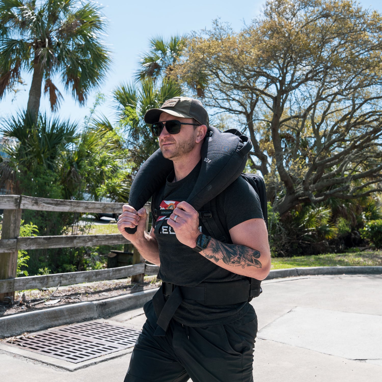 A man wearing sunglasses walks outside with a Bulgarian Sandbag over his shoulders, featuring reinforced handles, among palm trees in the sun.