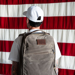 Wearing a white cap and shirt, a person with a large backpack featuring the "Patch - Embrace The Suck" stands in front of a U.S. flag.