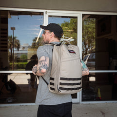 A man with tattoos and a cap stands outside a glass building on a sunny day, wearing the large beige GR1 backpack.