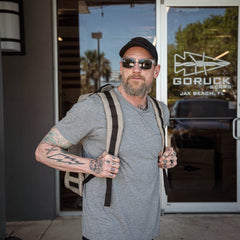 A tattooed man in sunglasses wears a GR1 backpack while standing outside a store with glass doors.