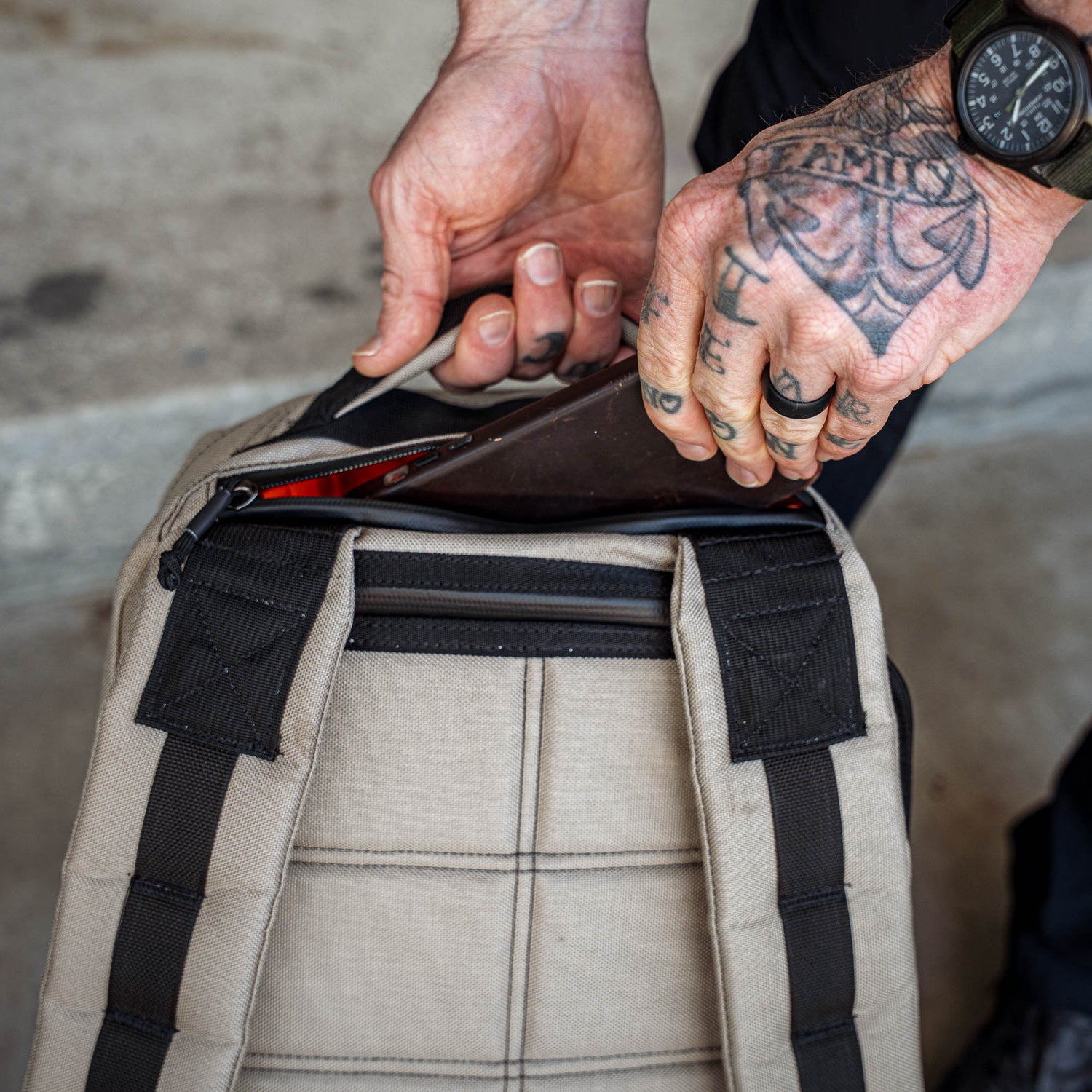 Tattooed hands place a tablet into a tan GR1 backpack with black straps, viewed from above—making the GR1 an ideal everyday carry companion.