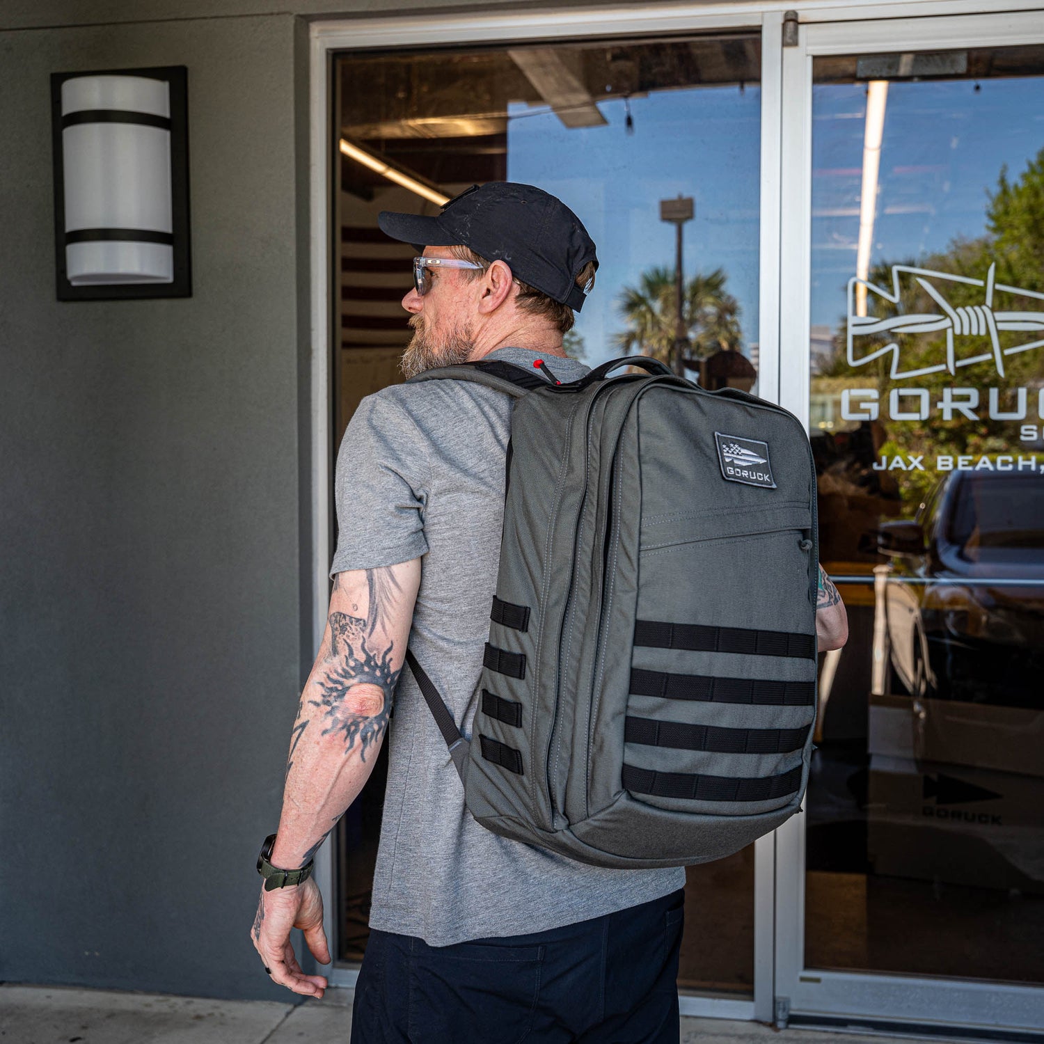 A man wearing sunglasses and carrying a GR2 travel backpack stands outside a building with glass doors displaying the 