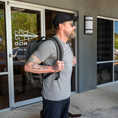 A man with tattoos, sunglasses, a cap, and a GR2 travel backpack stands outside a building with glass doors.