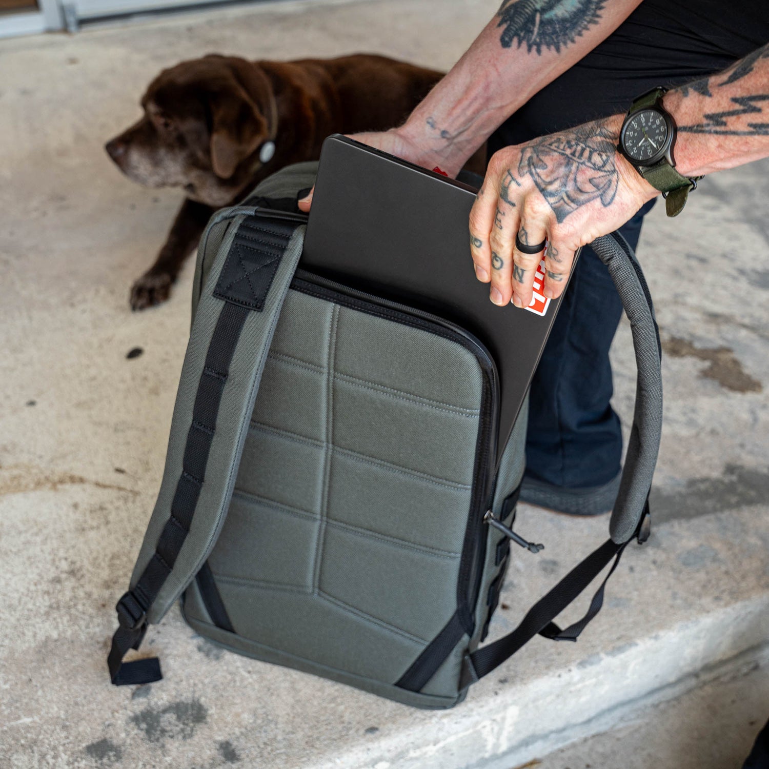 A tattooed person slips a laptop into a gray GR2 travel backpack, while a brown dog rests nearby on the ground.