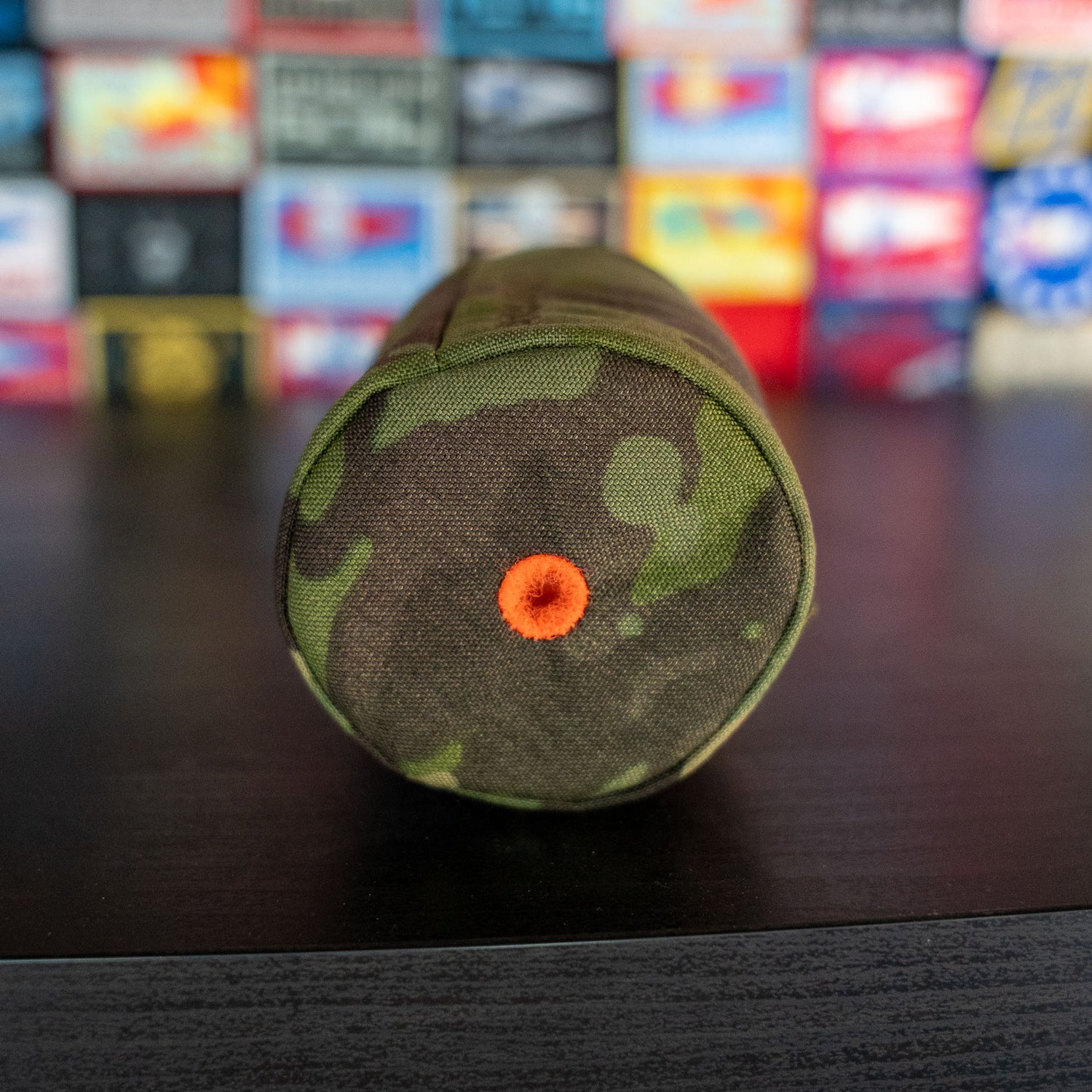 A Ballistic Beer Jacket, featuring a round, camouflage-patterned collapsible design with an orange circle in the center, stands upright on a dark surface.