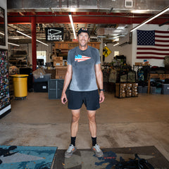 A man in athletic wear smiles in a warehouse with an American flag and gear behind him, wearing Men’s Training Shorts - ToughStretch, featuring sweat-wicking performance fabric.