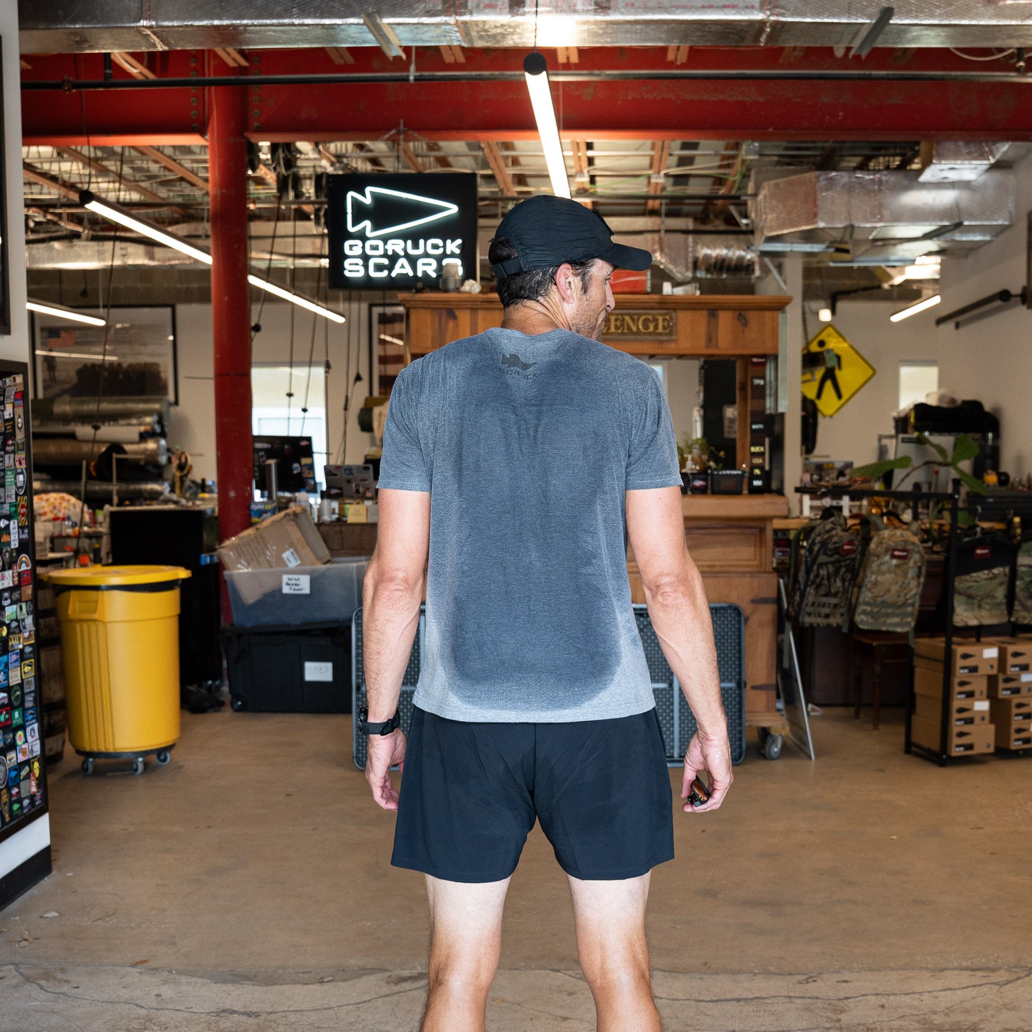 A man in athletic gear, wearing the GORUCK Flag Spearhead Tee - Unisex Tri-BlendX with a visible sweat stain and logo on his back, stands inside a gear store facing away from the camera.