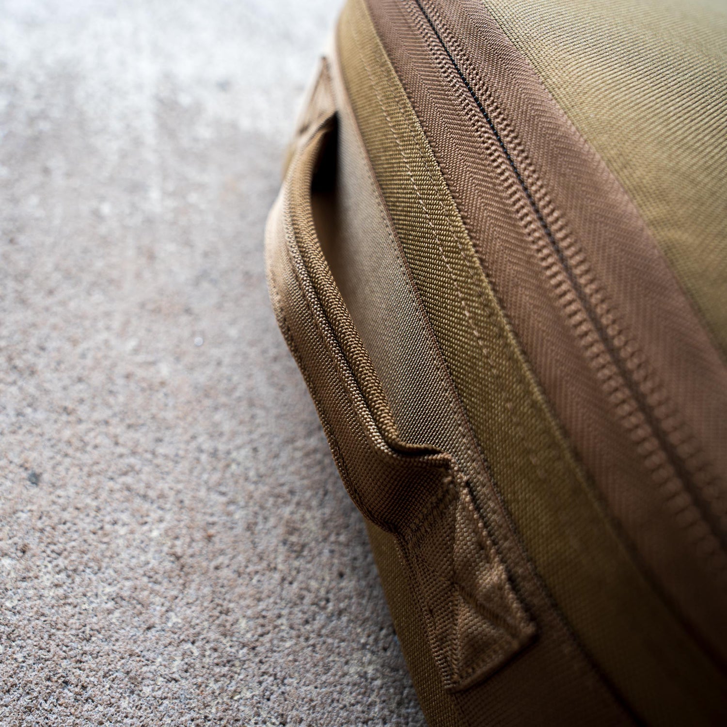 Close-up of a Rifle Case made from brown fabric, featuring a zipper and handle, resting on a textured concrete surface.