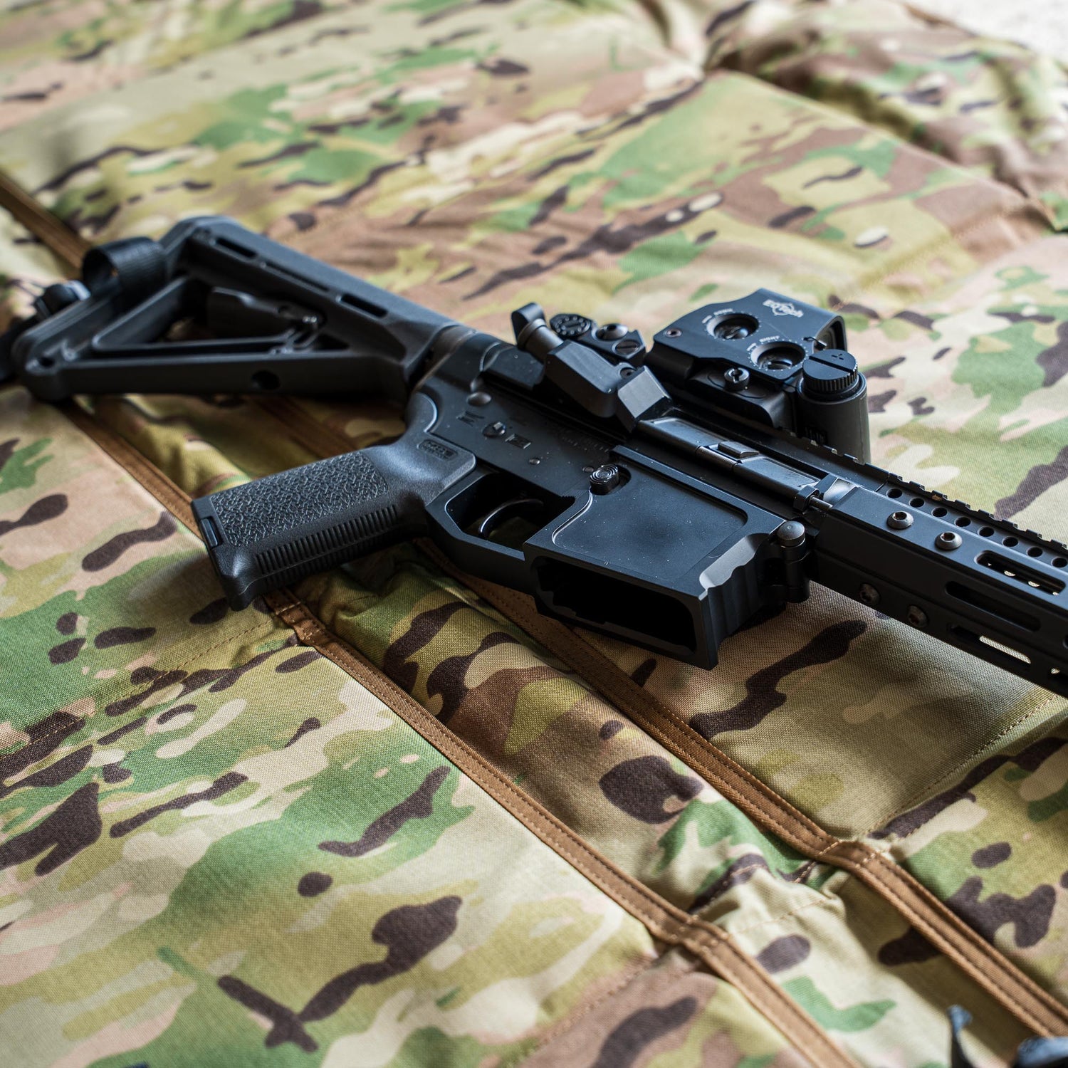 A black tactical rifle with a scope rests on the Shooting Mat, which features a camouflage pattern.