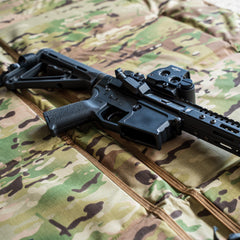 A black tactical rifle with a scope rests on the Shooting Mat, which features a camouflage pattern.