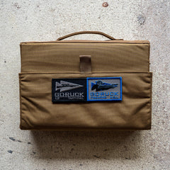 The Ammo Book is a brown tactical bag with two GORUCK patches, designed for ammo storage and shown on a textured, light-colored surface.