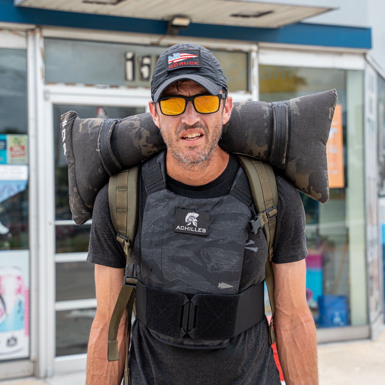 A man in the Achilles | Men's Weighted Vest and sunglasses carries a large camouflage pillow on his shoulders outside a building.