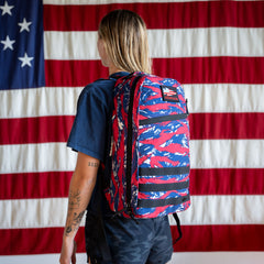 Someone with a Bullet Ruck Classic - Ballistic Nylon Cordura 15L daypack in red, white, and blue stands before an American flag.