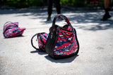 A Sand Kettlebells bag with "GORUCK" text and a padded handle, featuring a red, white, and blue pattern, rests on sunlit pavement—durable gear you can count on.