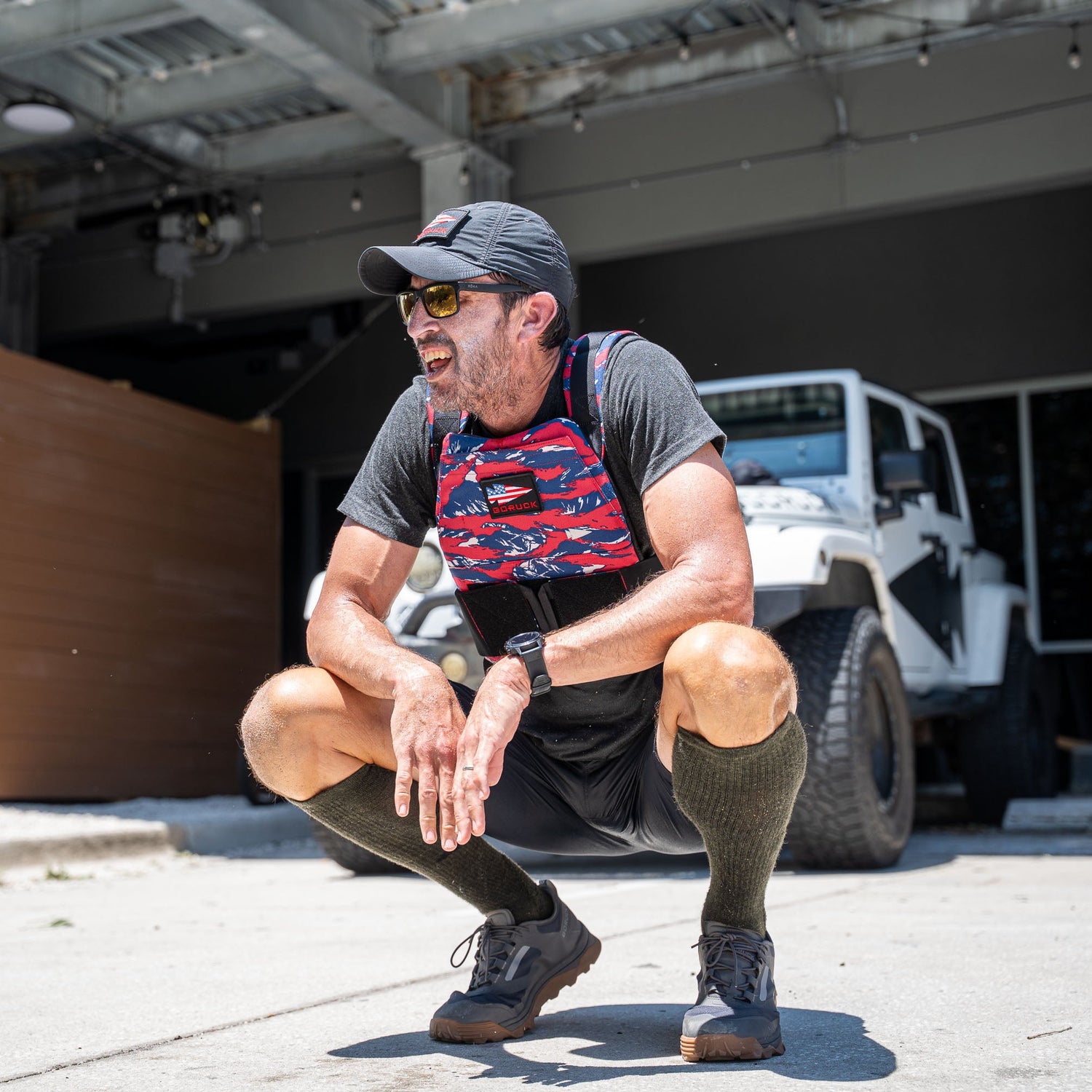 A man squats outdoors in athletic gear and a Rucking Weight Vest, with a white jeep and garage behind him—ideal for rucking sessions or training using curved ruck plates.