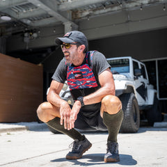A man squats outdoors in athletic gear and a Rucking Weight Vest, with a white jeep and garage behind him—ideal for rucking sessions or training using curved ruck plates.