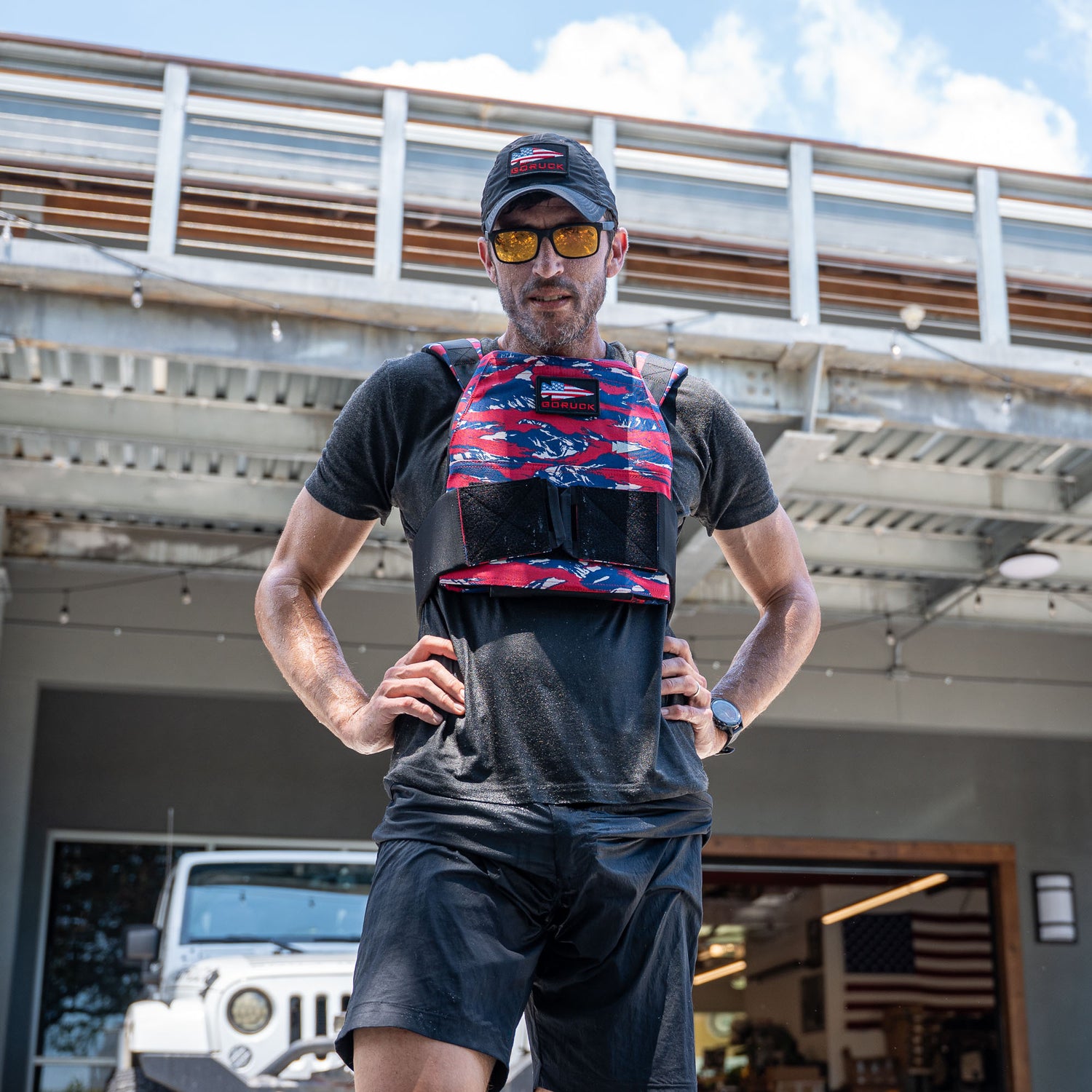 Man in black shorts and t-shirt wears a Rucking Weight Vest with curved plates, plus sunglasses and cap, standing outdoors ready for rucking.