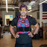 Man wearing sunglasses, a hat, and the Rucking Weight Vest in red, white, and blue stands indoors with American flags in the background—ready to ruck in patriotic style.