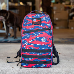 A Bullet Ruck Classic - Ballistic Nylon Cordura backpack in red, blue, and white rests on the ground with a blurred dog and boxes in the background.