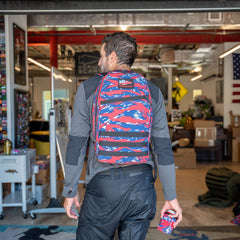 A man carrying a colorful Bullet Ruck Classic - Ballistic Nylon Cordura 15L backpack and matching item enters a workspace lined with boxes and patriotic decorations.