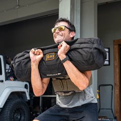 Wearing sunglasses and lifting a weighted sandbag outdoors, this man is all smiles as he enjoys his rucking workout—made even more intense with the Rucking Weight Vest.