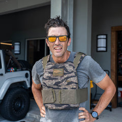 A man stands outside near a garage and a white vehicle, looking at the camera while wearing sunglasses and the Rucking Weight Vest—ready for some serious rucking.
