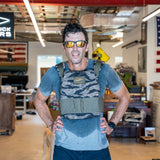 A man wearing sunglasses and the Rucking Weight Vest stands sweating in a cluttered indoor space with flags and signs, likely after an intense rucking session.