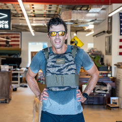 A man wearing sunglasses and the Rucking Weight Vest stands sweating in a cluttered indoor space with flags and signs, likely after an intense rucking session.