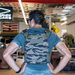 A man stands with hands on hips in a workshop, seen from behind, wearing a Rucking Weight Vest with ruck plates, ready for a rucking session.