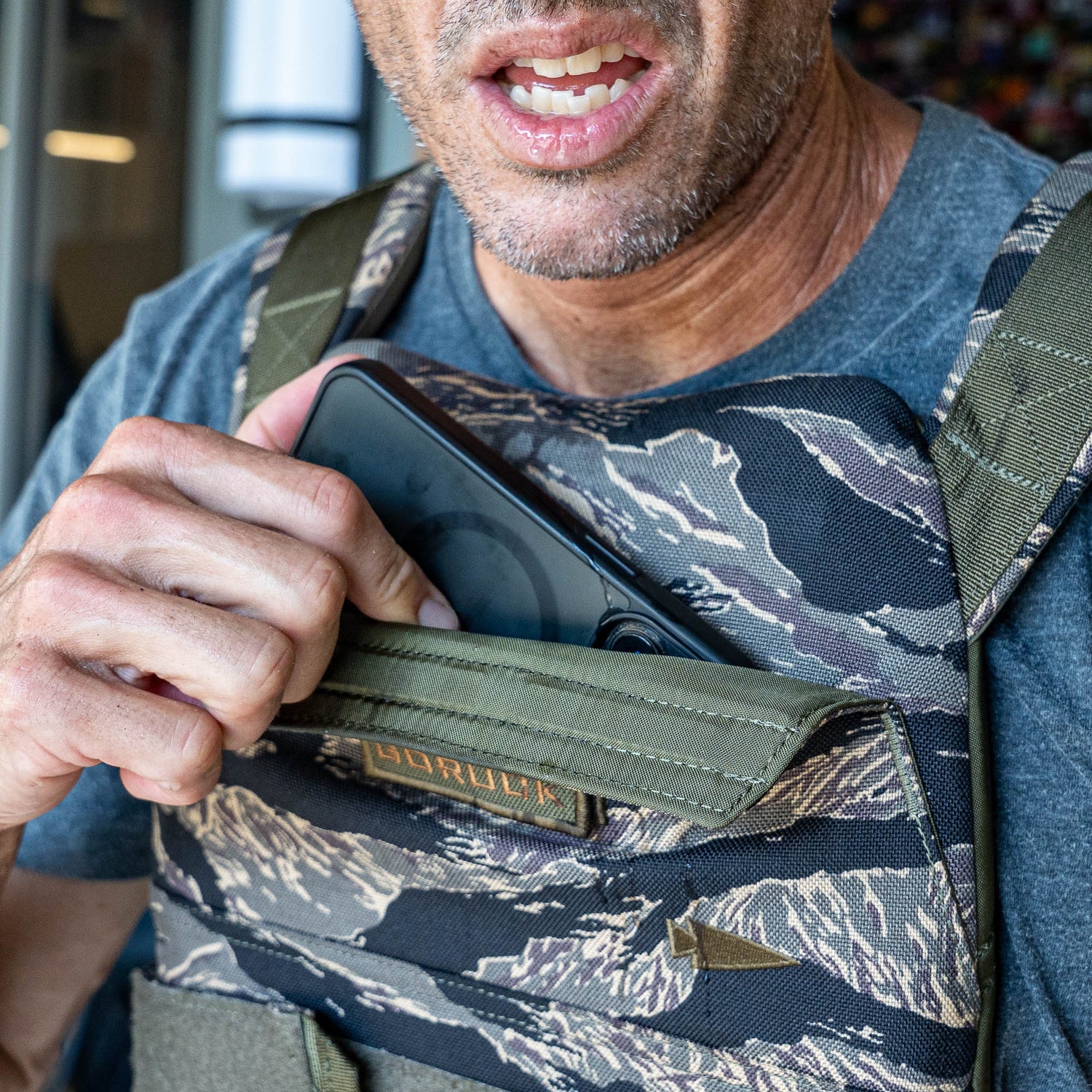 A man places a phone into the front pocket of the Rucking Weight Vest, which features a camouflage pattern, his mouth slightly open.