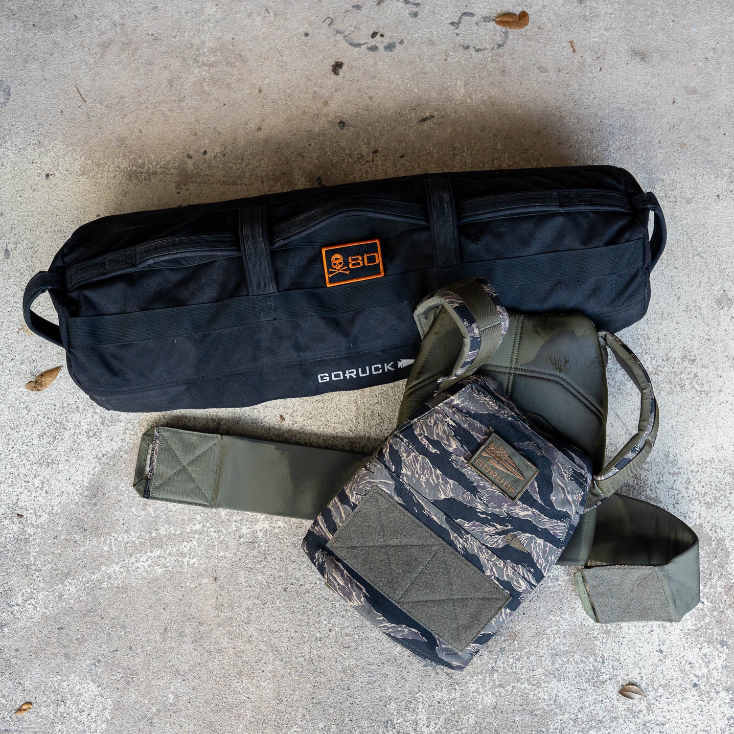 A Rucking Weight Vest with a camouflage pattern and a black duffel bag rest on a concrete floor scattered with leaves, both ready for you to add your ruck plates for the next challenge.