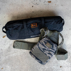 A Rucking Weight Vest with a camouflage pattern and a black duffel bag rest on a concrete floor scattered with leaves, both ready for you to add your ruck plates for the next challenge.
