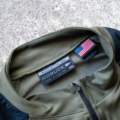 Close-up of the Warm Up Full Zip jacket collar in olive green, featuring visible GORUCK and USA flag labels that highlight its ToughDry shoulders. This lightweight jacket is proudly made in the USA.