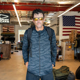 Man in sunglasses and a dark, lightweight Warm Up Full Zip stands indoors with an American flag and gear bags behind him. The jacket features ToughDry shoulders and is proudly made in USA.