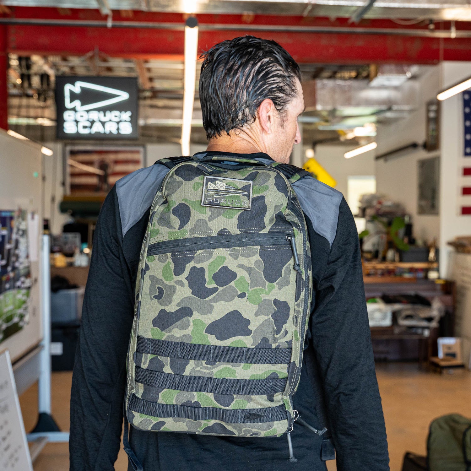A man wearing the Bullet Ruck Classic - Ballistic Nylon Cordura backpack stands indoors facing away, with workshop equipment and signs visible in the background.