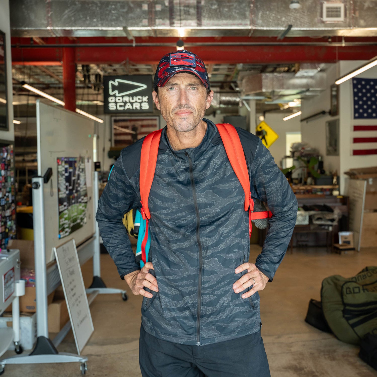 A man wearing a cap and the Mesh Ruck quick-drying backpack stands in an industrial room with an American flag on the wall.